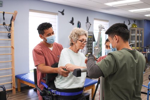 Two therapists helping patient with standing balance using support harness in rehab gym