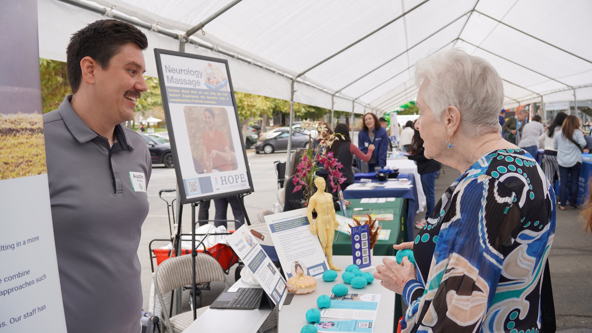 HOPE Clinic staff member explaining neurology massage services to elderly visitor at booth